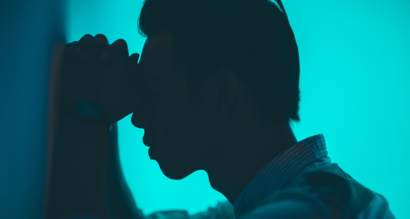 A silhouette of man against a teal background, with hands on forehead, leaning against a wall.