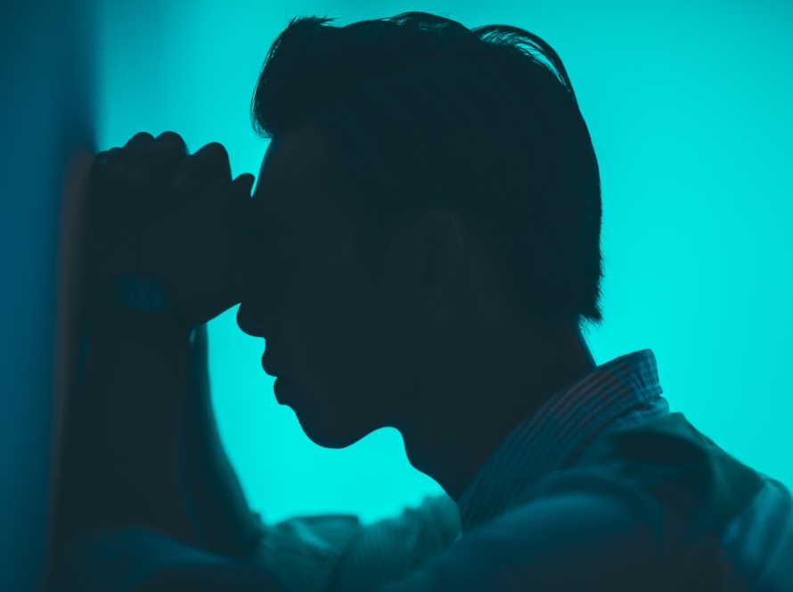 A silhouette of man against a teal background, with hands on forehead, leaning against a wall.