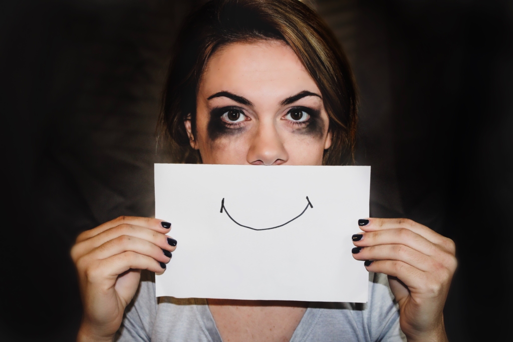 An image of a woman hiding behind a paper with a fake smile, here mascara is smudged, indicating she's just been crying