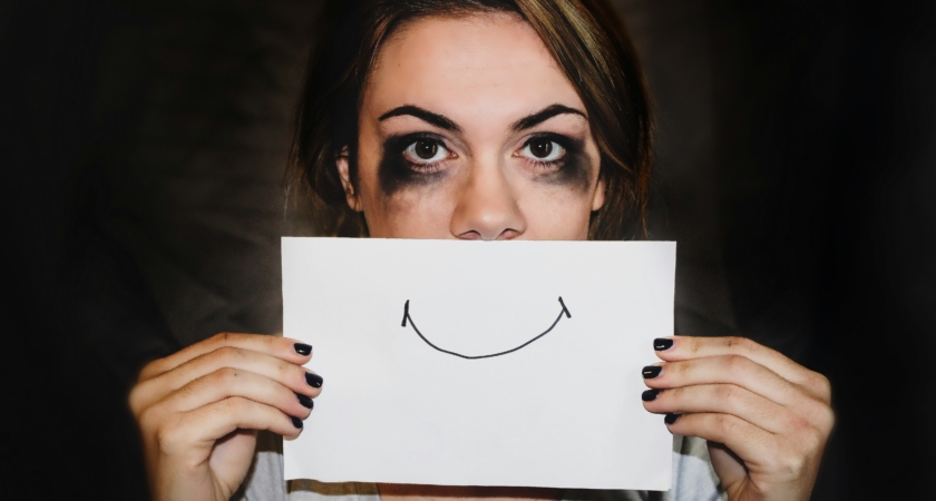 Woman with smudged mascara around eyes, holding up a paper with a smiley drawn on it.