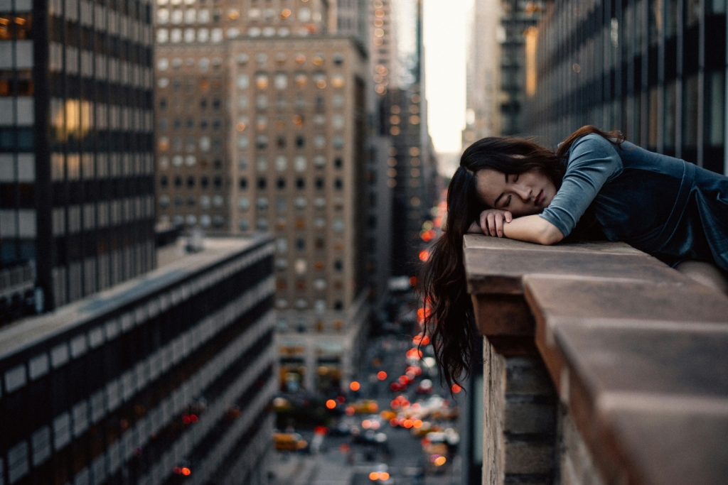 Woman tired out leaning over a building with traffic beneath looking visibly suffering from burnout